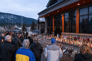 A quiet Canadian mountain town gathers in mourning outside a civic building with candles, flowers, and soft dusk ligh