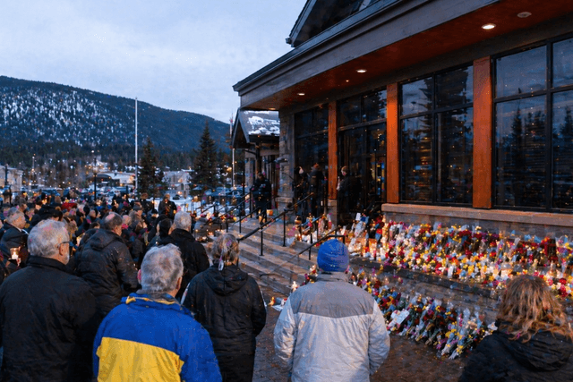 A quiet Canadian mountain town gathers in mourning outside a civic building with candles, flowers, and soft dusk ligh