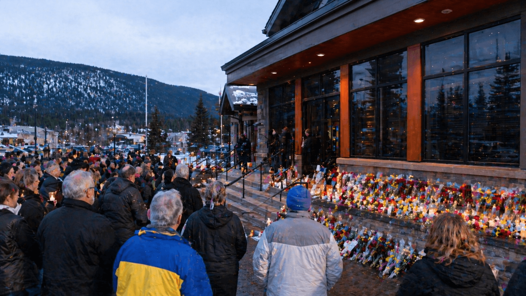 A quiet Canadian mountain town gathers in mourning outside a civic building with candles, flowers, and soft dusk ligh