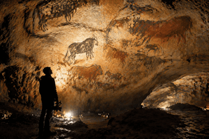 A cinematic view inside an ancient cave with prehistoric paintings glowing under soft torchlight, while a lone modern figure stands observing the artwork.